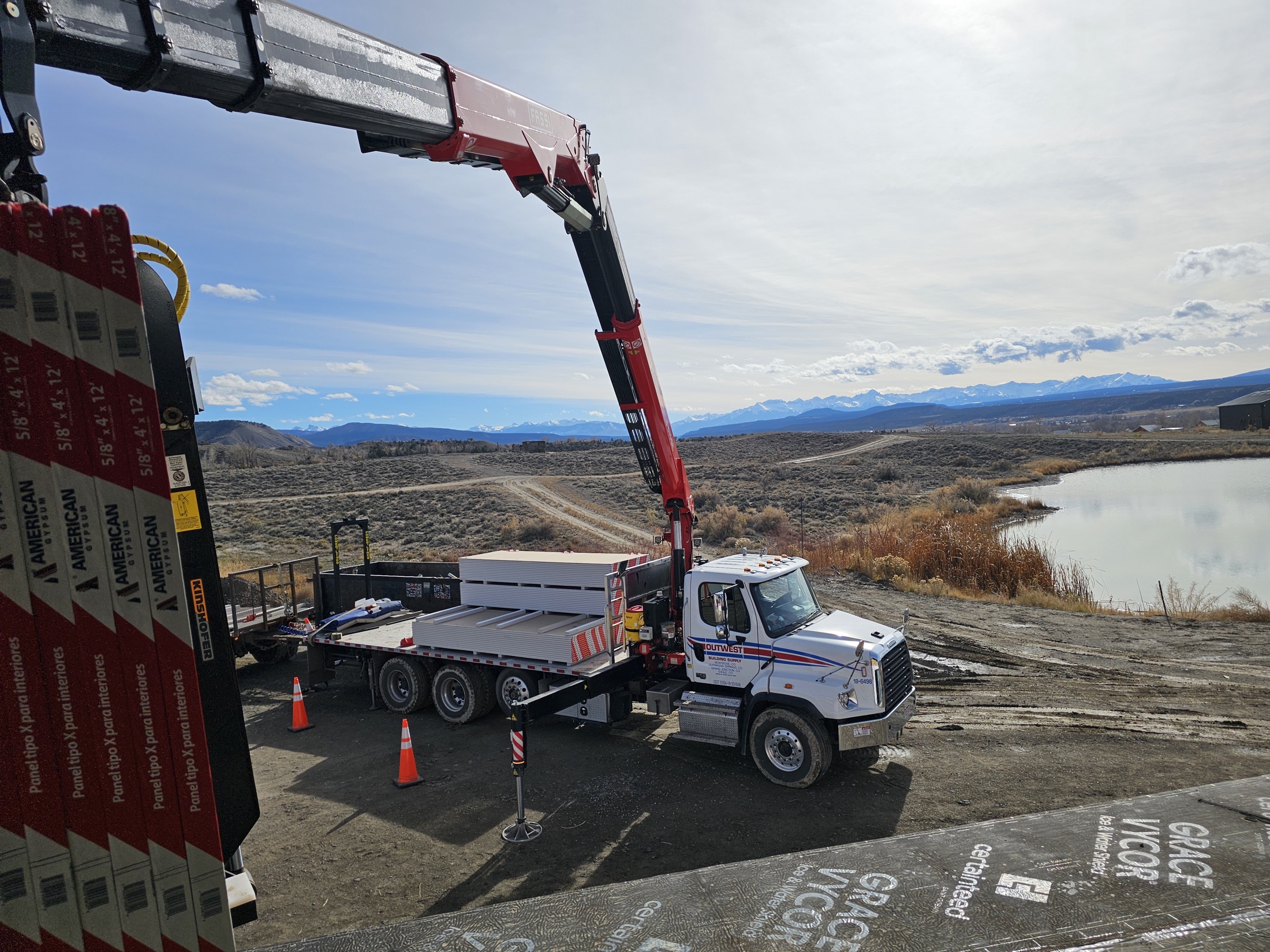 Outwest boom truck unloading drywall at a jobsite beside a lake and mountains